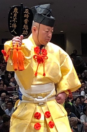 Photograph of sumo umpire Kimura Yōdō in yellow outfit, with calligraphy-bearing gunbai raised.